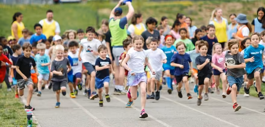 Dozens of kids sprinting off the start line on a track, volunteers in pinnies cheering behind them